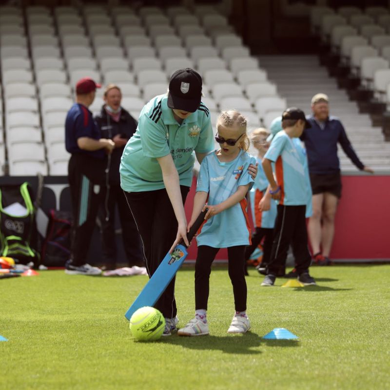 A lady guiding a young girl with a vision impairment to hit the large cricket ball with a bat.