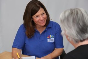 Image shows an Eye Clinic Liaison Officer (ELCO) in a branded Sight for Surrey top with a pen in her hand resting on top of a piece of paper on a table, looking the person infront of her.