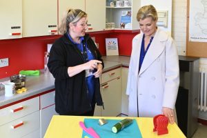 The Duchess & Deanne Weller in a kitchen setting. A table setting is in front of them with a bright yellow table cloth. On the top is a blue chopping board, pink knife, red vegetable holder and a finger guard.