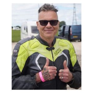 Nigel Lamb, aka Blind Bloke Racing. Nigel is smiling directly at the camera holding two thumbs up, he is wearing motorcycle leathers and dark glasses.