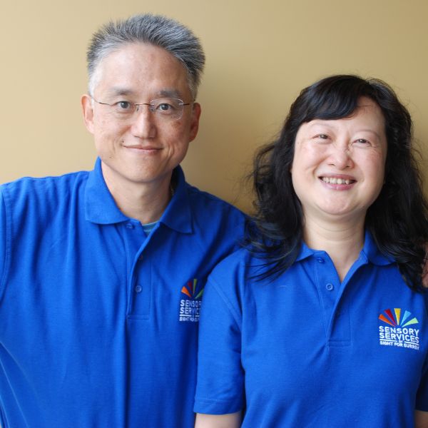 Image shows volunteers Patrick and Susan standing against a wall, both are wearing Sight for Surrey blue tops