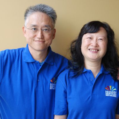 Image shows Patrick and Susan, Home Visitor Volunteers for Sensory Services by Sight for Surrey. Both are wearing blue t-shirts with the charity's logo and the couple are standing against a coloured wall smiling at the camera.