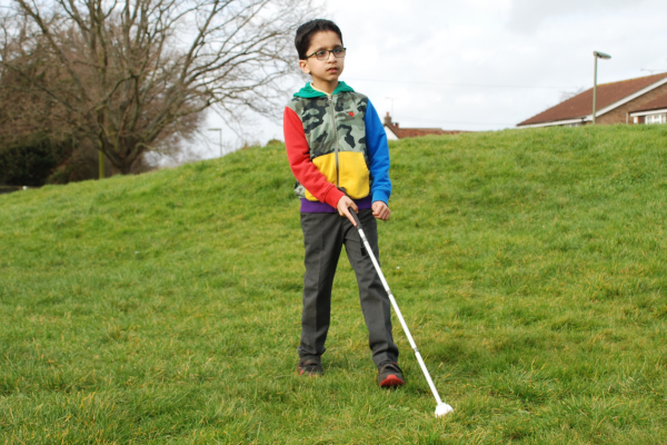 Image shows a young boy wearing a colourful top and grey trousers walking on grass using a white cane.