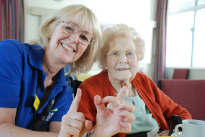 Image shows, Katie from the charity sitting alongside an elderly lady they are both smiling at the camera and making the sign 101 with their fingers.