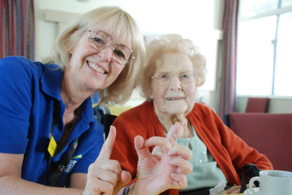 Image shows, Katie from the charity sitting alongside an elderly lady they are both smiling at the camera and making the sign 101 with their fingers.