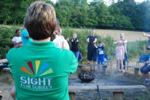 The back of a person wearing a Sight for Surrey t-shirt facing a group of adults and children around a campfire.