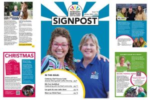 Image shows the front cover of Signpost that features celebrity Chef, Yvonne Cobb and Emma Burton from the charity. Around the front cover are other pages from the magazine giving people an insight into the content of this edition of Signpost.