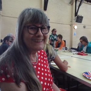 Image shows a lady smiling at the camera, she is seated at a table and behind her in the background are other people.