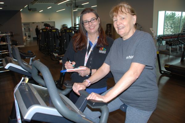 Image shows a lady at the gym on an exercise bike. Standing next to her is a member of staff from the charity, she is writing in a notebook.