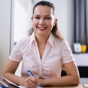 Image shows a young lady smiling at the camera, she has her arms resting on the desk infront of her and is holding a pen in one hand.