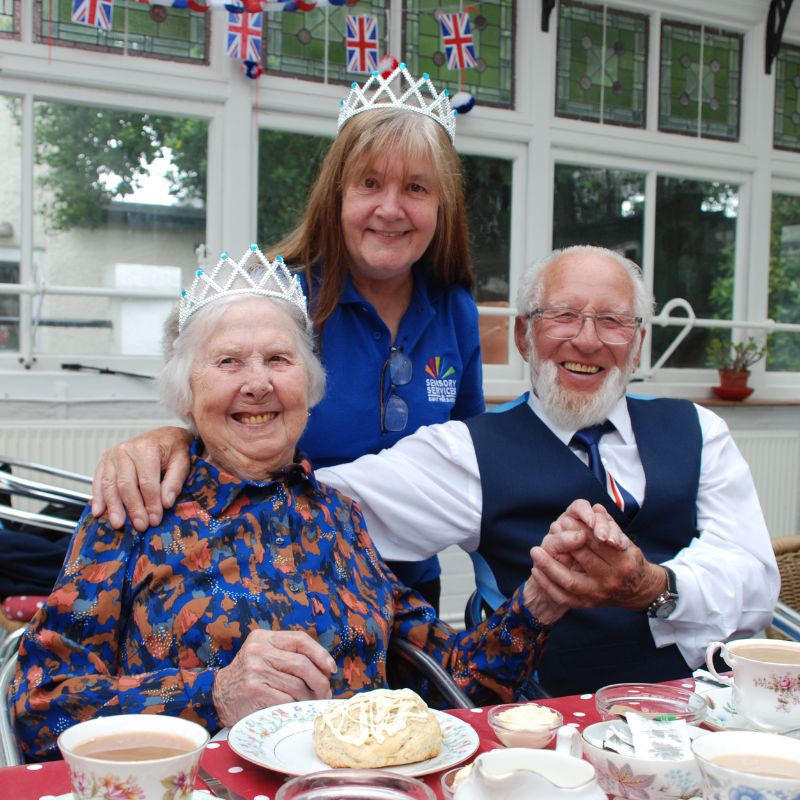Image shows Julie Barclay standing behind two people we support who are sitting at a table laid out with delicious looking scones, jam and cups of tea.