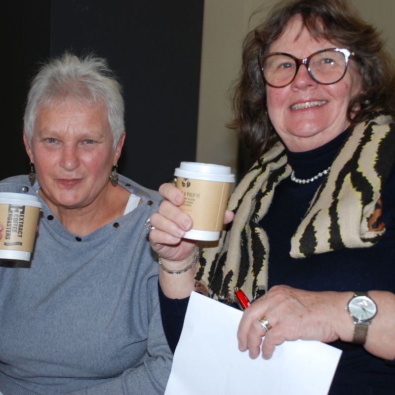 Two ladies sitting side by side both are drinking from coffee cups and smiling at the camera