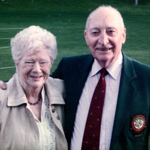 Image shows an elderly couple standing next to each other smiling, the man is wearing a suit and tie and has one around the lady with his hand just showing on her shoulder.