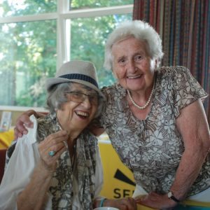 Image features two ladies, one standing up with her arm around the shoulders of the other lady sitting down. both are smiling into the camera.