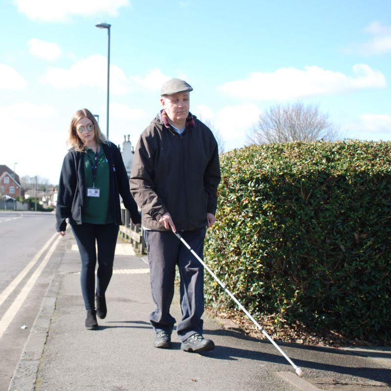Image shows a man walking with a white cane along a pavement a member of staff from the charity walks behind him looking over his shoulder, they are next to a road with double yellow lines running into the background