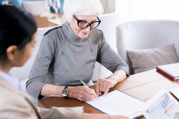 Image shows an older lady sitting at a desk writing on a piece of paper, another lady is looking over her with her hand at the top of the piece of paper.