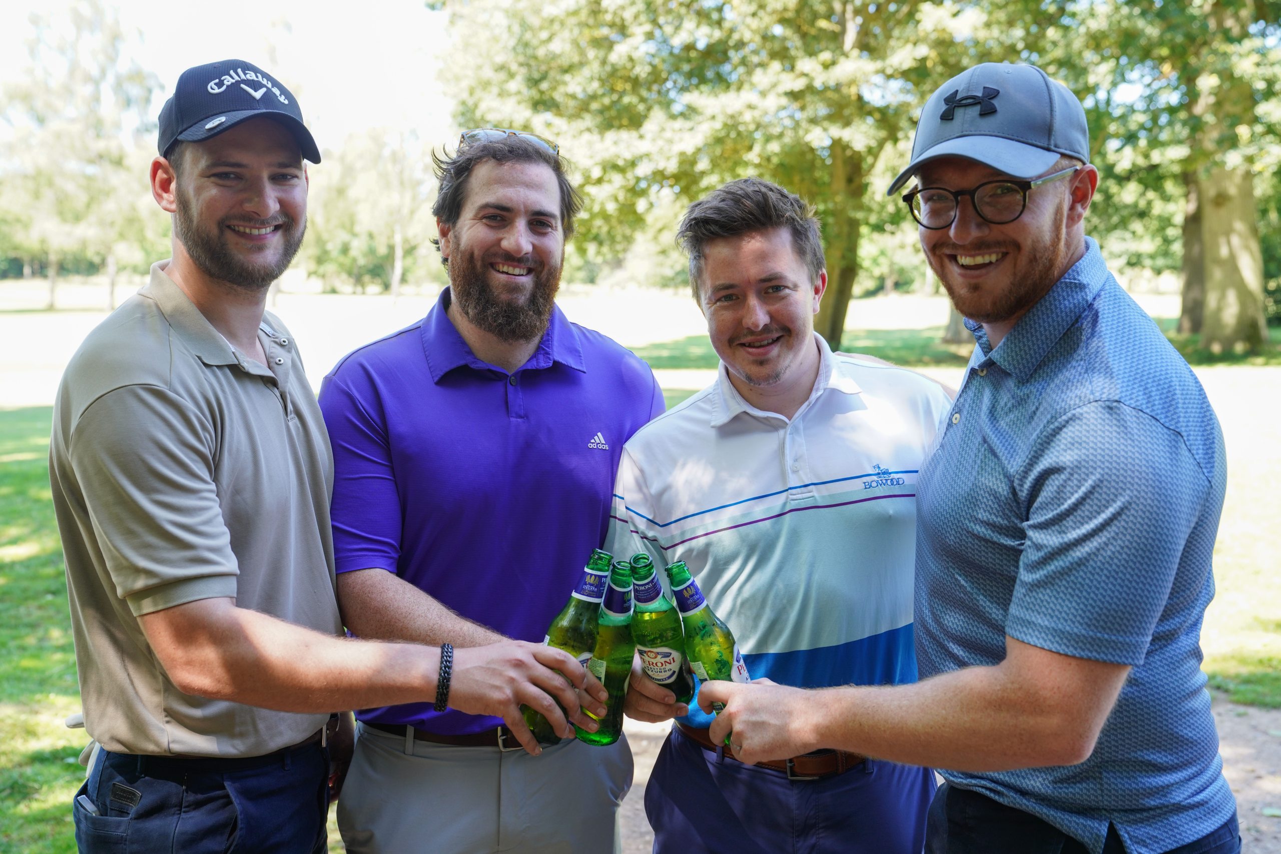 Image shows a group of men outside on a golf course they are all holding a bottled drink and clinking their bottles together. They are all smiling at the camera.
