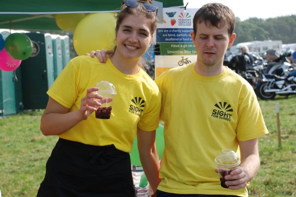 Image shows a young woman and man wearing Sight for Surrey t-shirts. The woman is holding a cup in one hand and smiling at the camera. A busy event can be seen in the background.