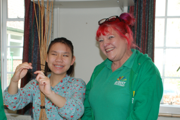 Image shows a member of staff with a young girl they are willow weaving, both are beaming at the camera.