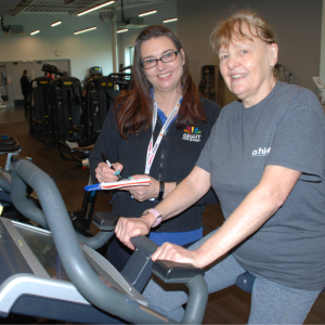 Image shows Suzy, a Communicator Guide, standing next to Larraine who is sitting on an exercise bike in a gym setting. 