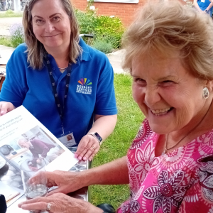 Image shows a lady with a beaming smile and a member of staff from the charity is sitting to her side, there is some literature infront of them.