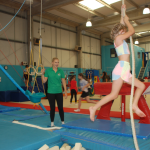 Image shows: a young girl swinging from a rope hanging down from the ceiling, a member of staff from the charity is standing nearby watching, there are other children and young people taking part in gymnastics behind her.