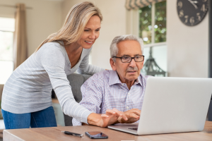 Image shows an elderly map at his laptop with a younger lady leaning over his shoulder helping him on his laptop.