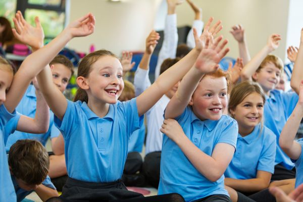 Image shows a group of young children sitting on the floor they are all raising their hands in the air.