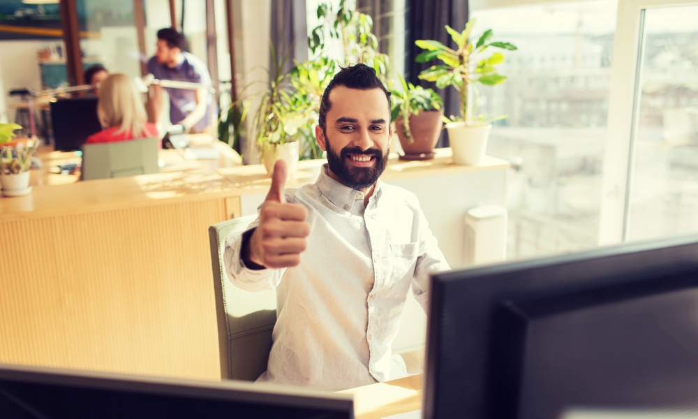 Man sits at desk in office giving merry thumbs up. He is surrounded by computer screens and colleagues are behind him.