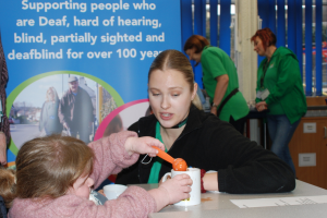 Image shows a member of staff from the charity kneeling at a table helping a young woman to measure and pour ingredients into a mug.