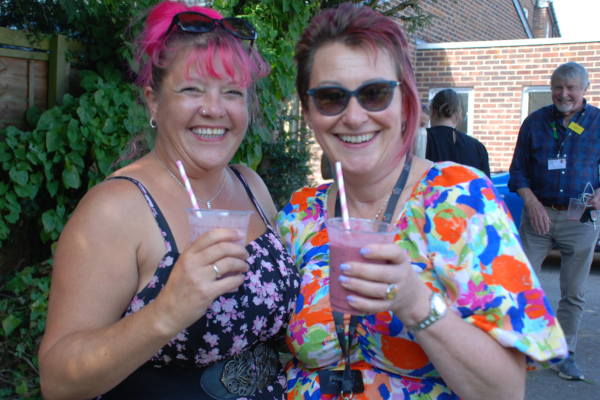 Image shows two members of staff from the charity drinking delicious smoothie drinks they have blended themselves on our smoothie bike, they are both holding cups with their blended smoothies and are smiling at the camera.