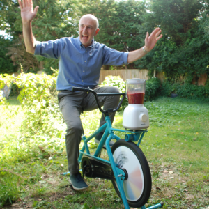 Image shows Tony Greene, Resource Centre Manager pedalling on our smoothie bike with both of his arms raised in the air.