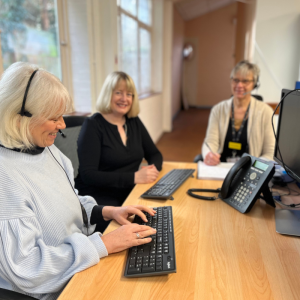 Image shows three ladies sitting around a desk wearing headsets, a keyboard and screen are on the desk.