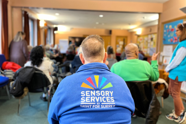 Image shows a member of staff from Sensory Services by Sight for Surrey at the Deaf Cafe in Redhill, he has his back to the camera and a sea of people can be seen infront of him.