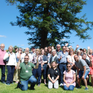 A large group of people from our Deaf Cafe in Redhill are standing outside in a park, the sky is blue and there is large tree behind them. The group are waving hello.