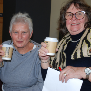 Two attendees to the Deaf Cafe are sitting holding coffee cups and smiling at the camera.