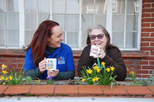 Two women are standing outside the charity offices in Fetcham. They are both holding a mug of tea and are sharing a joke as both are laughing.