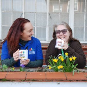 Two ladies are standing next to each other both are holding branded Sensory Services by Sight for Surrey mugs. They are both laughing and having fun. 