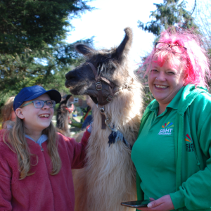 Fi from our CYPS Team is standing next to a llama. A young girl is the other side and they are both laughing and smiling.