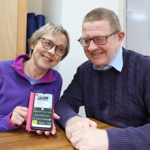 The photo shows, Gary, Tech Trainer with a lady we support, Gary is showing her some accessibility features on an iPhone. The lady is holding the iPhone is her hands and is smiling at the camera.