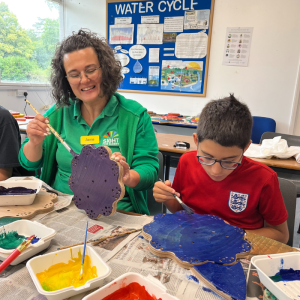 A member of staff from the Children and Young People's Team is sitting at a table with a young boy, they are painting a wooden structure.