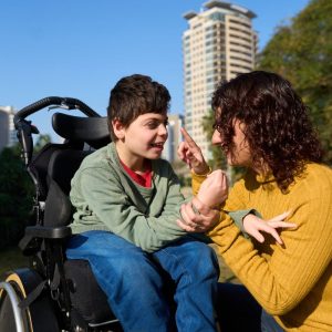 A young boy is sitting in a wheelchair and a lady is crouched down beside him