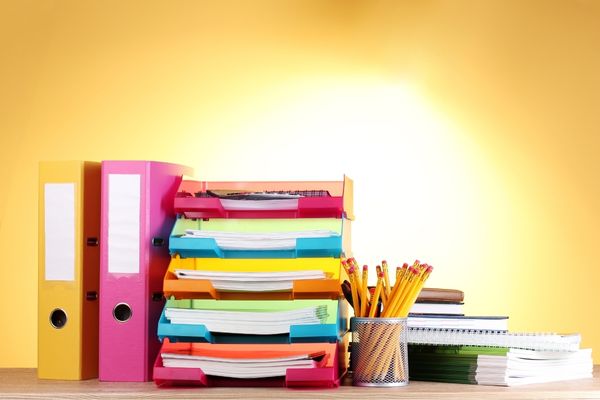 A number of brightly coloured files are stacked up on a desk alongside a pot of pens.