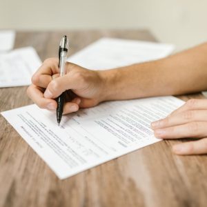 A person is filling in a form on a table, you can see their hand is holding a pen and part of their arm is visible.