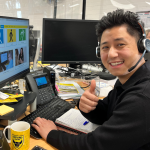 Helpdesk Advisor Hang is at his desk in front of phone and two computer monitors giving a thumbs up to the camera, wearing a phone headset.