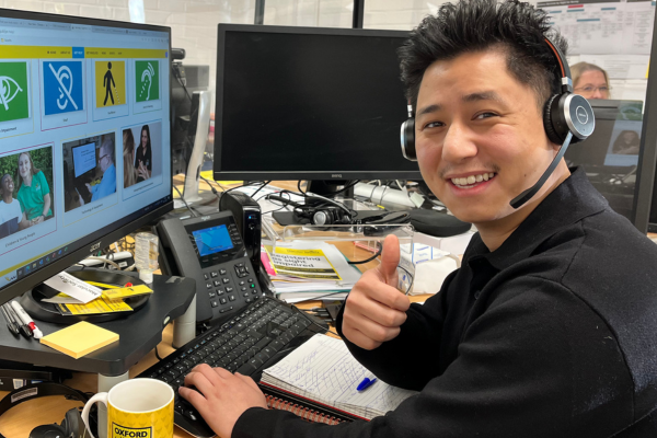 Helpdesk Advisor Hang is at his desk in front of phone and two computer monitors giving a thumbs up to the camera, wearing a phone headset.