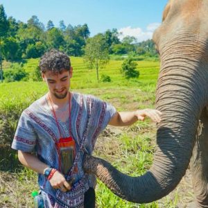 Calum is standing next to an elephant. He has one arm reaching out to touch the elephant.