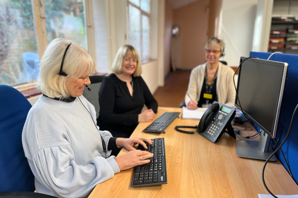 A group of three woman are sitting around a desk, one has a telephone headset on and is typing on a keyboard