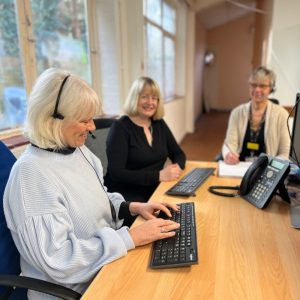 Three woman are sat around a desk with keyboards and telephone headsets on.