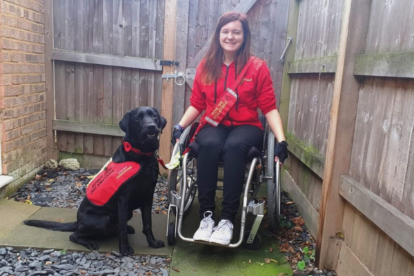 Megan is sitting in a wheelchair, she is wearing a red top and black leggings, sitting next to her is her Assistance Dog, it is a black Labrador.